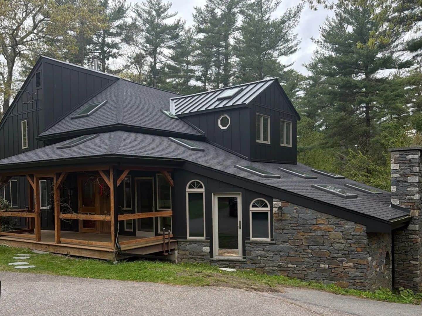 Modern farmhouse in Carver, MA with new black board-and-batten siding, a black shingle roof with skylights, a timber-frame porch, and a stone wall