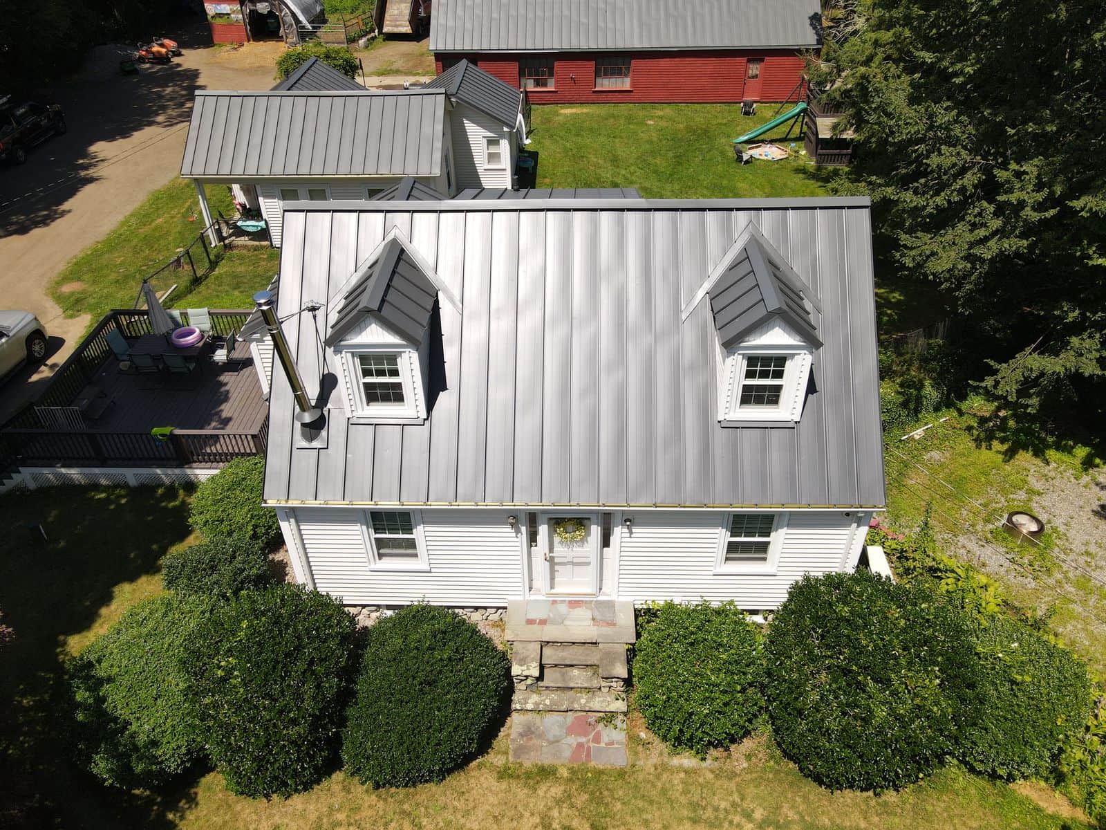 Aerial view of a white cape in Rehoboth, MA with a new gray standing-seam metal roof and two dormers, the red barn with its matching metal roof behind