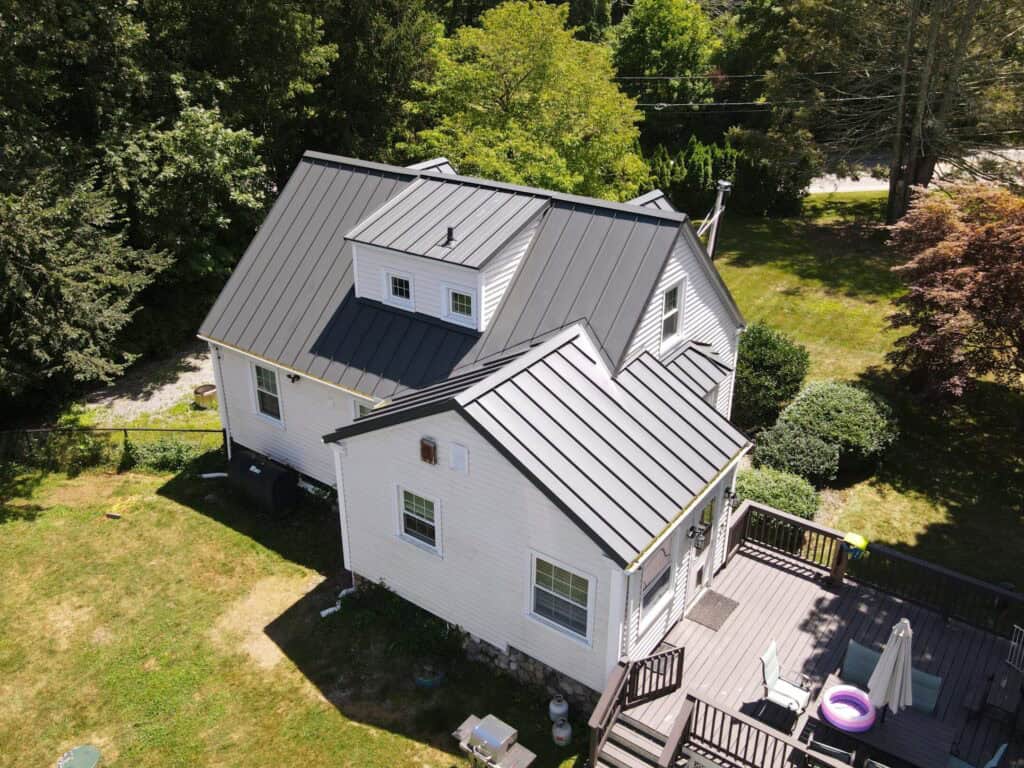 Metal roof on a white residential house with a spacious backyard, surrounded by green trees, featuring a deck with outdoor furniture and an umbrella.
