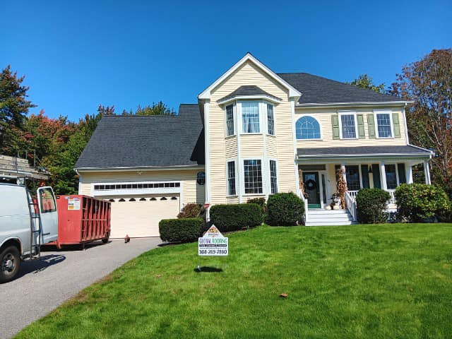 Roof of a modern two-story house with a dark asphalt shingle roof, beige exterior siding, and well-maintained front yard.
