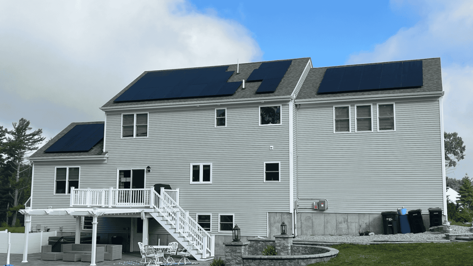A large two-story New England home with light gray siding and multiple sections of sleek black solar panels installed across the roof, maximizing sun exposure for energy savings.