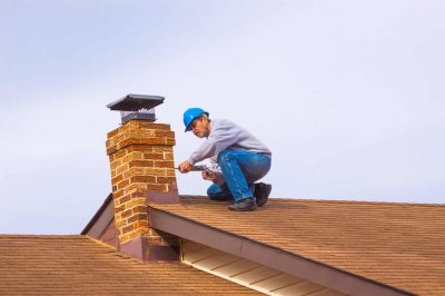 Chimney inspection and maintenance worker on roof, with tools, working on brick chimney with chimney cap.