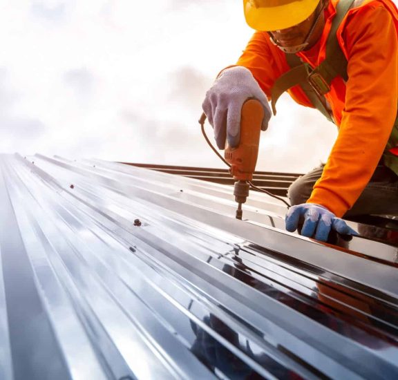 Shingling specialist installing or repairing a galvanized metal roof, wearing safety gear and using a power drill.