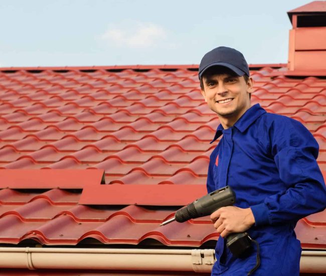 Young happy man contractor worker in blue overalls is repairing a red roof with electric screw driver. He is smiling and looking to the camera. Roofing concept. Copy space for you text
