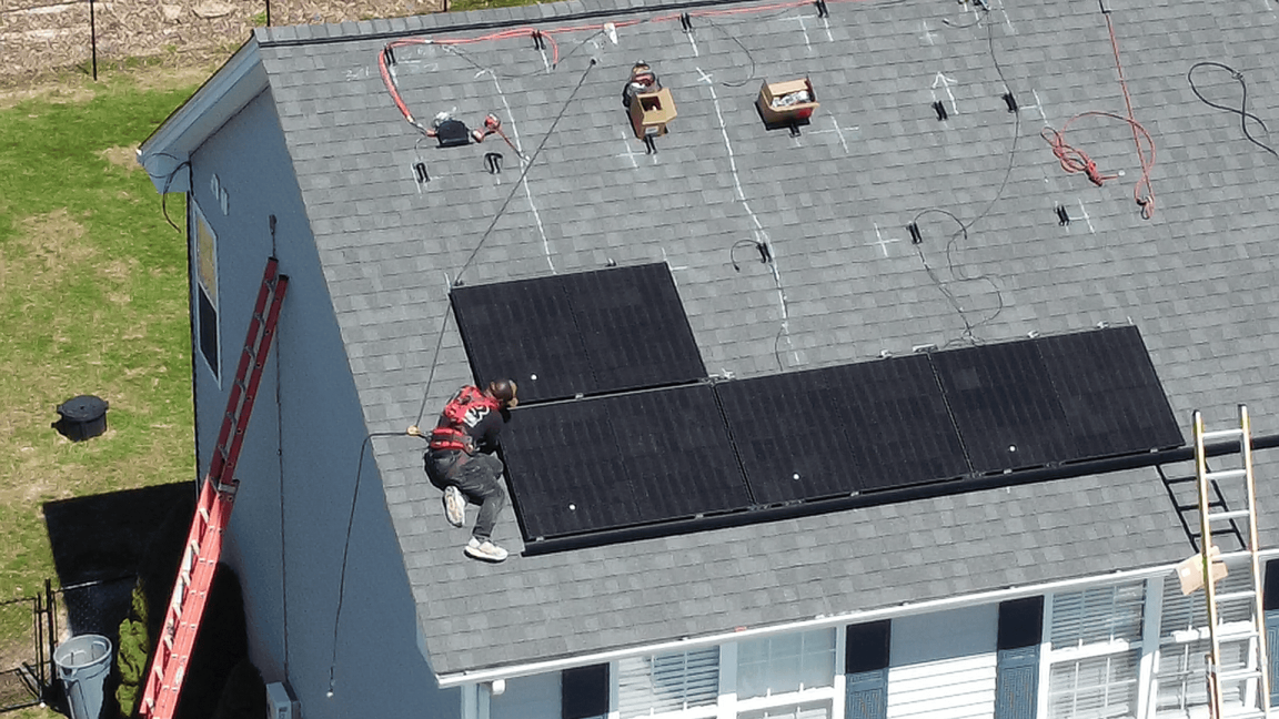 A New England home with a gray shingle roof during an in-progress solar installation. A worker in safety gear is securing black solar panels while tools, wiring, and equipment are set up across the roof.