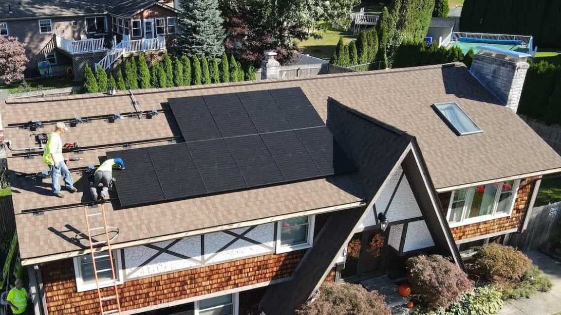 A New England home with a brown shingle roof where solar panels are being installed. Two workers in safety gear are actively securing panels while ladders and equipment are set up for the in-progress project.